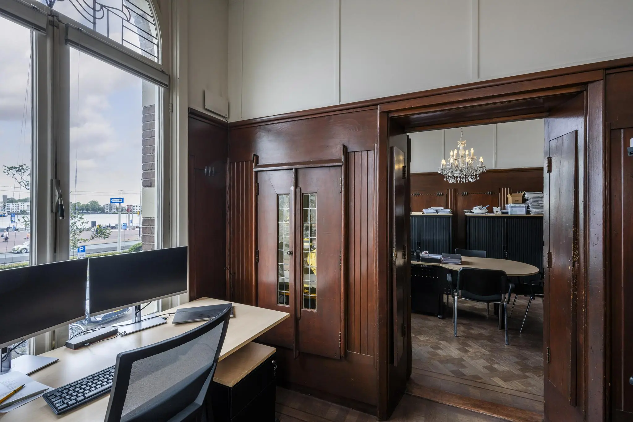 Kantoorruimte aan de De Ruijterkade met houten afwerking, werkplek bij het raam en doorkijk naar vergaderruimte met kroonluchter.